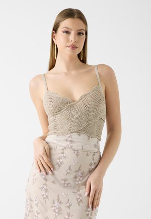 Ung kvinde med glat hår iført guldringe, en beige strikket cropped top med tynde stropper og et beige blomsterbroderet skørt.