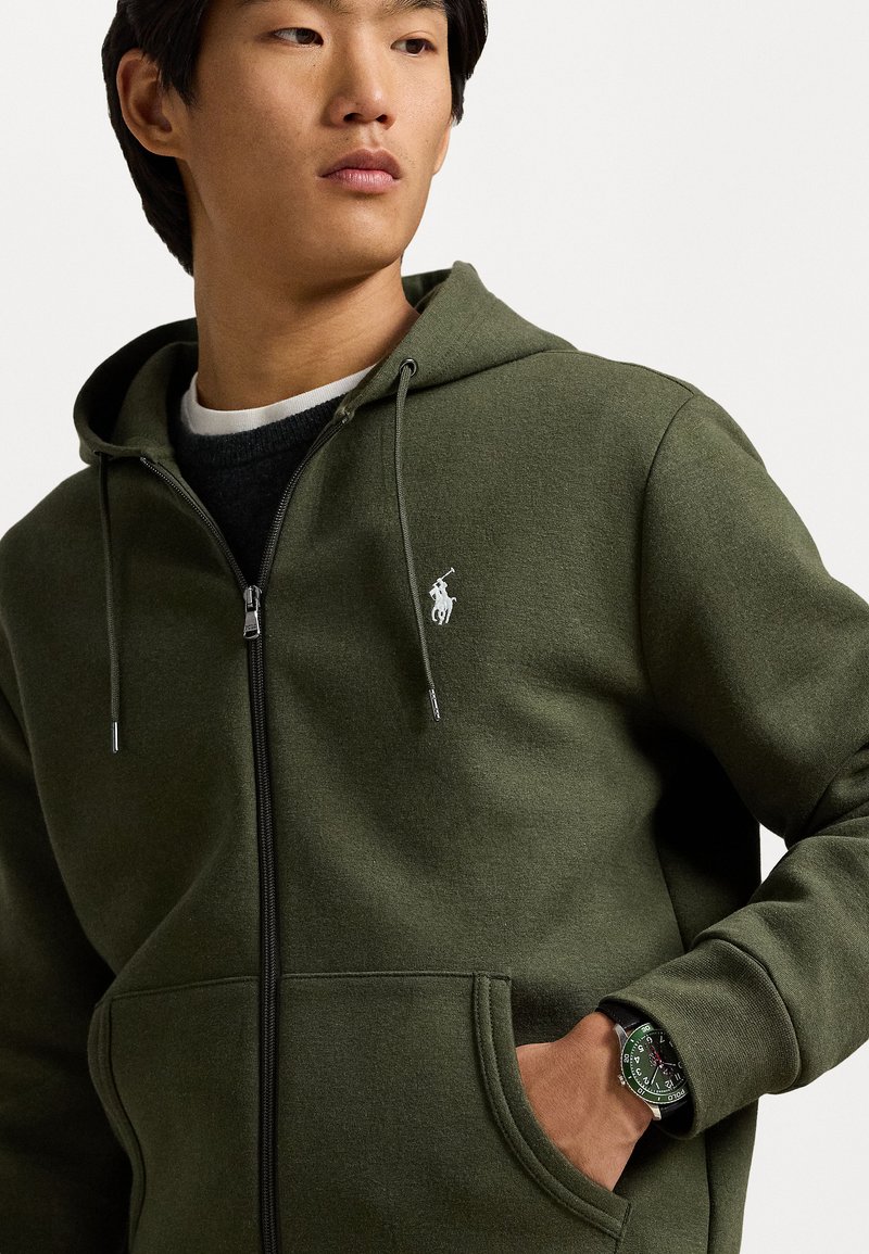 Sudadera con cremallera en verde olivo, fabricada en un tejido suave, con un pequeño logo blanco en el lado izquierdo del pecho, bolsillos laterales y capucha con cordón.