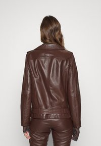 Donna con lunghi capelli castani che indossa una giacca di pelle marrone scuro e pantaloni abbinati, vista da dietro su uno sfondo neutro.