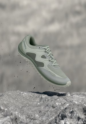 Zapatilla de correr Merrell color verde claro suspendida en el aire sobre un terreno rocoso con partículas de tierra dispersas alrededor.
