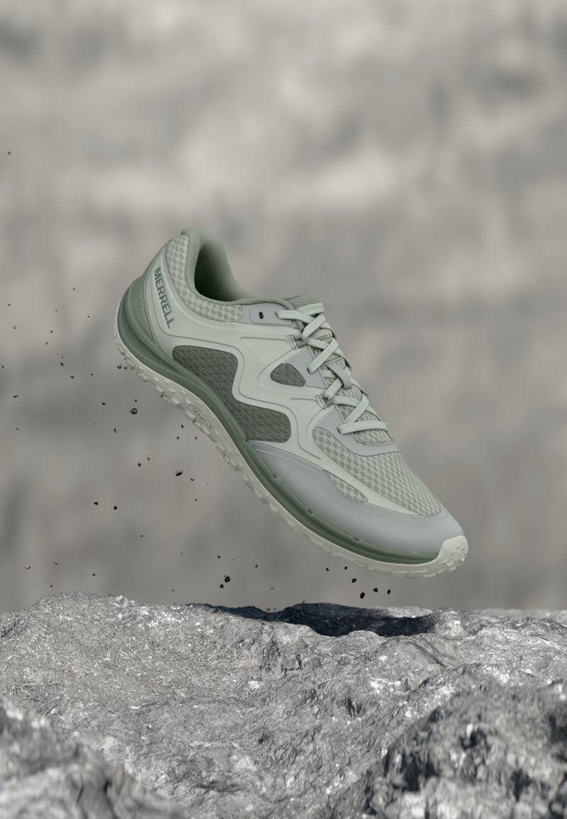 Zapatilla de correr Merrell color verde claro suspendida en el aire sobre un terreno rocoso con partículas de tierra dispersas alrededor.