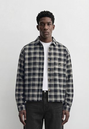 Mand iført en sort- og hvidternet skjorte med knapper over en hvid T-shirt og sorte bukser, stående foran en ensfarvet grå baggrund.