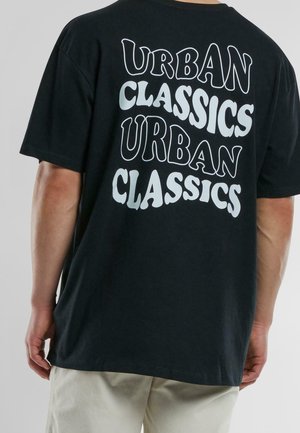 Person iført en sort T-shirt med "URBAN CLASSICS" trykt to gange i store, hvide, stiliserede bogstaver på ryggen.