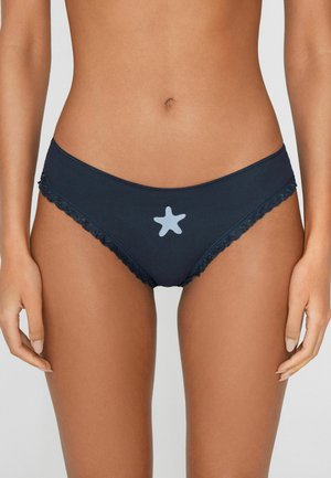 Kobieta w granatowym bieliźnie w stylu bikini z jasnoniebieskim wzorem gwiazdy i koronkową ozdobą na jednolitym tle.