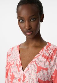 Korallenmuster-Bluse mit V-Ausschnitt und dezenten Falten. Sie hat eine glatte Textur und unregelmäßige weiße abstrakte Designs.
