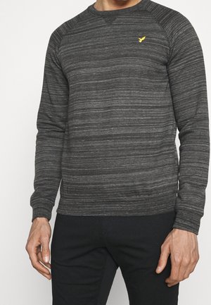 Homme portant un pull rayé gris foncé à manches longues avec un petit logo jaune d’oiseau sur la poitrine, associé à un pantalon noir, debout sur un fond blanc.