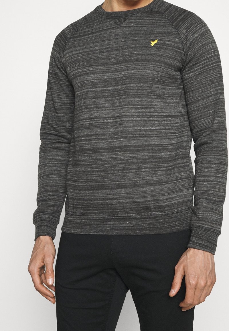 Homme portant un pull rayé gris foncé à manches longues avec un petit logo jaune d’oiseau sur la poitrine, associé à un pantalon noir, debout sur un fond blanc.