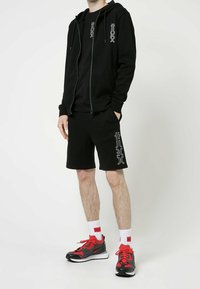 Felpa nera con zip, t-shirt nera e pantaloni corti neri con dettagli grafici bianchi. Abbinati a scarpe da ginnastica rosse e nere e calzini bianchi.