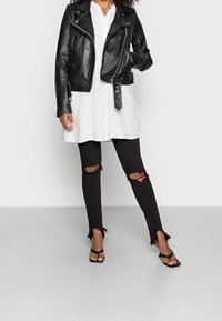 Chaqueta de motorista de cuero negro sobre un vestido túnica blanco y leggings negros rasgados, con dobladillos deshilachados y sandalias negras de tacón.