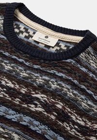 Strikket sweater med rund hals, der har et flerfarvet mønster i mørkebrun, blå og creme farver. Mærket siger "anerkjendt."