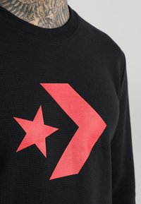 Nahaufnahme eines schwarzen strukturierten Shirts mit einem großen roten Stern und einem Wellenmuster auf der Brust, das einen tätowierten Hals über dem Kragen zeigt.
