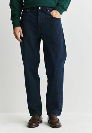 Man wearing dark blue straight-leg jeans, green sweatshirt, and brown leather loafers, standing with one hand in pocket.