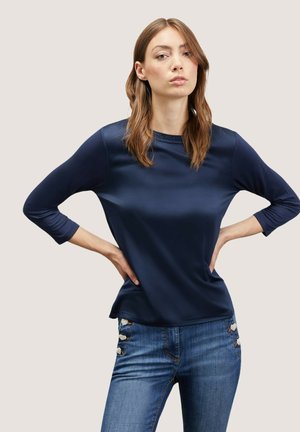 Donkerblauwe top met lange mouwen in een gladde stof en een ronde halslijn. Gecombineerd met denim jeans met decoratieve hardware op de zakken.
