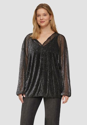 Blonde Frau, die eine schwarze transparente Bluse mit langen Ärmeln und metallischen Fäden sowie dunkelgraue Jeans trägt, vor einem schlichten Hintergrund stehend.