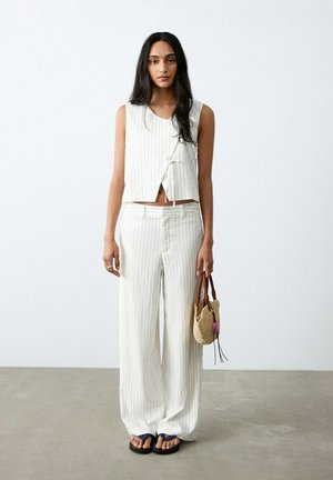 Mujer con cabello largo y oscuro, vestida con un top sin mangas blanco con rayas finas y pantalones anchos a juego, sosteniendo un pequeño bolso tejido.