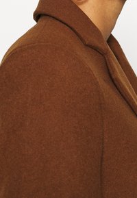 Brun uldfrakke med en struktureret krave, der har en tekstureret overflade og synlige syningsdetaljer. Sidevinklen fremhæver det skræddersyede design.