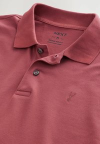 Camisa polo color burdeos de tela texturada, con cuello, dos botones y un pequeño logotipo bordado en el pecho. Diseño de corte regular.