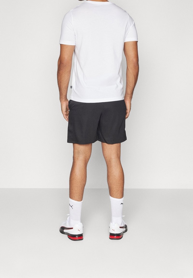 Hombre de pie de espaldas, vistiendo una camiseta blanca, pantalones cortos negros, calcetines blancos de Puma y zapatillas blancas con acentos rojos.