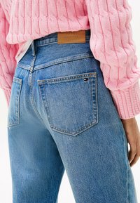 Jeans de tiro alto, de mezclilla azul claro, con dos bolsillos traseros. Presenta una etiqueta de cuero marrón con "TOMMY HILFIGER" en relieve.