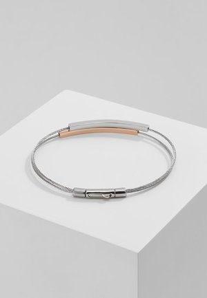 Bracelet fin en métal tressé argenté avec une barre incurvée bicolore et un petit fermoir en métal sur une plateforme blanche.