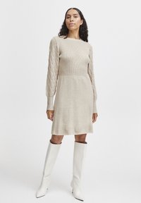 Strickkleid in hellem Beige mit geripptem Oberteil, kabelgestrickten Ärmeln und einem ausgestellt geschnittenen Rock, kombiniert mit strukturierten weißen kniehohen Stiefeln.