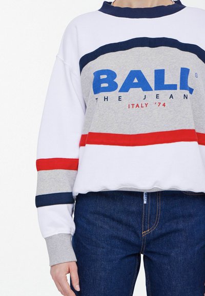 Sweatshirt blanc et gris avec des rayures rouges et bleues, arborant le logo 'BALL THE JEAN ITALY '74'. Associe à un jean en denim bleu foncé.