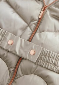 Puffer bunda vo svetlo béžovom saténe, s ružovo-zlatým zipsom, cvokmi a elastickým pásom s prešitým vzorom.