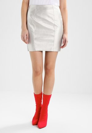 Femme portant une mini-jupe argentée brillante et des bottines chaussettes à talons hauts rouge vif, debout sur un fond uni.