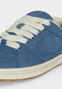 Baskets en daim bleu avec lacets blancs épais, panneau latéral perforé, semelle intermédiaire blanche et semelle extérieure en caoutchouc naturel sur fond clair.