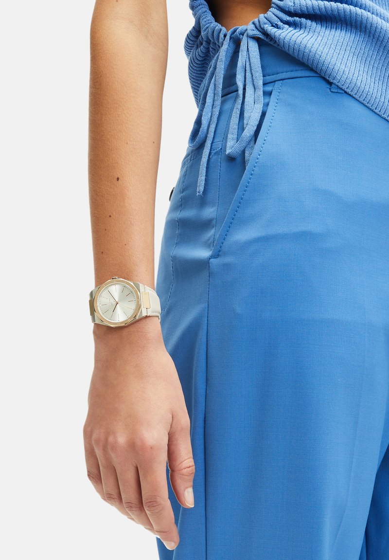 Zilveren polshorloge met een ronde witte wijzerplaat en een minimalistisch ontwerp, gecombineerd met lichtblauwe stoffen broek en een bijpassend blauw topje.