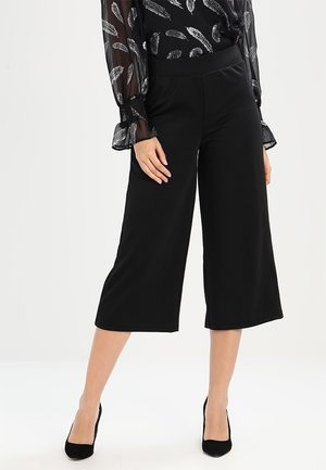 Femme portant un pantalon noir large et court, un chemisier noir transparent avec des motifs de plumes blanches, et des escarpins noirs à bout pointu, debout avec la main sur la cuisse.