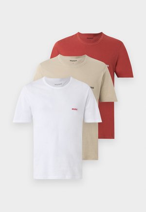 Homme debout pieds nus, portant une chemise blanche à manches courtes avec le logo "HUGO" et un short bleu marine ajusté sur un fond clair uni.