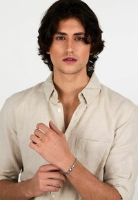 Bracciale d'argento al polso, caratterizzato da un design a maglie. Presenta una finitura lucida ed è indossato sopra una camicia di lino beige chiaro.