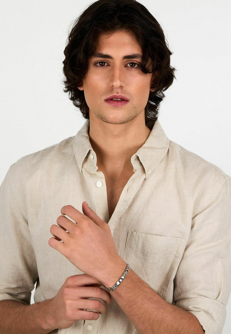 Bracciale d'argento al polso, caratterizzato da un design a maglie. Presenta una finitura lucida ed è indossato sopra una camicia di lino beige chiaro.