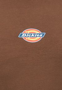 Καφέ ύφασμα με ένα μικρό λογότυπο Dickies στο κέντρο που εμφανίζει έναν στυλιζαρισμένο πορτοκαλί πέταλο και λευκό κείμενο σε μπλε λωρίδα.