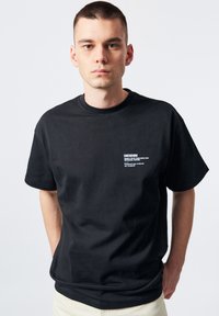 Camiseta de algodón negra con cuello redondo y mangas cortas. Presenta un logo impreso en blanco en el área del lado izquierdo del pecho. Diseño sencillo.