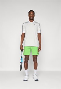 Graues Polohemd mit kurzen Ärmeln und navyblauen Akzenten, kombiniert mit knallgrünen Shorts. Weiße Socken und Tennisschuhe. Hält einen Tennisschläger.