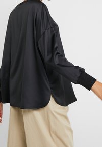 Person som bär en löst sittande svart satinblus med lång ärm och ribbade manschetter tillsammans med beige vida byxor, visad från ryggen och sidan.