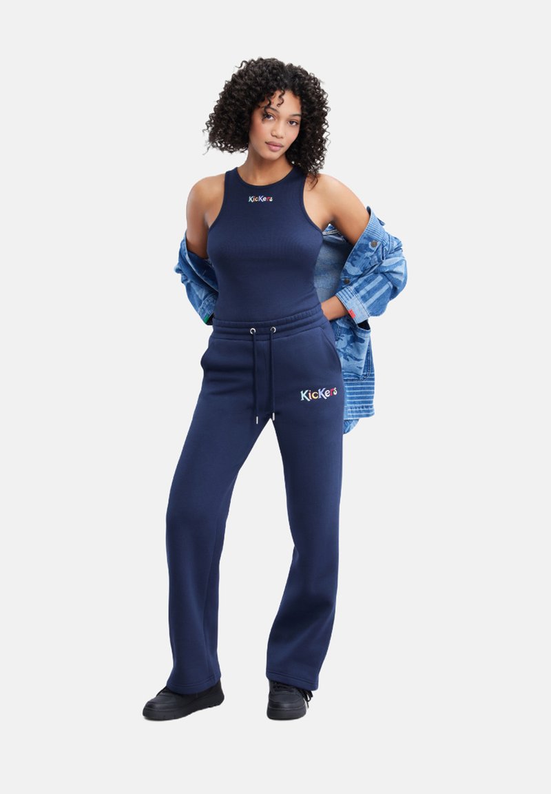 Canottiera navy e pantaloni della tuta, entrambi in cotone. I pantaloni della tuta presentano un cordino e dettagli con logo. Sovrapposti con una giacca di denim blu. Scarpe nere.