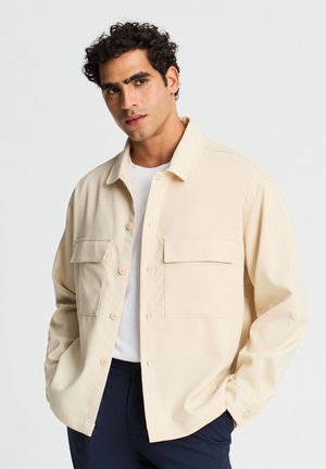 Uomo con i capelli ricci che indossa una camicia beige con bottoni sopra una t-shirt bianca e pantaloni scuri, in piedi con le mani in tasca.