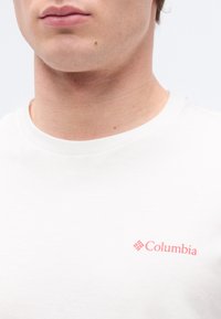 Κοντινό πλάνο ενός ατόμου που φοράει μια λευκή μπλούζα με στρογγυλή λαιμόκοψη και ένα μικρό κόκκινο λογότυπο Columbia στο στήθος.