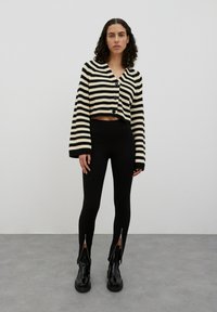 Zwart en crèmekleurig gestreepte cropped cardigan met knopen, gecombineerd met zwarte leggings met ritsdetails bij de enkel en glanzende zwarte laarzen.