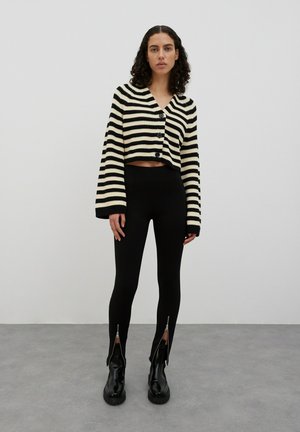 Zwart en crèmekleurig gestreepte cropped cardigan met knopen, gecombineerd met zwarte leggings met ritsdetails bij de enkel en glanzende zwarte laarzen.