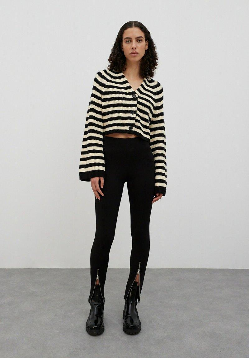 Zwart en crèmekleurig gestreepte cropped cardigan met knopen, gecombineerd met zwarte leggings met ritsdetails bij de enkel en glanzende zwarte laarzen.