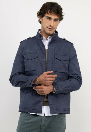 Hombre con cabello oscuro y rizado que lleva una chaqueta azul marino sobre una camisa blanca y pantalones verdes, de pie frente a un fondo blanco liso.