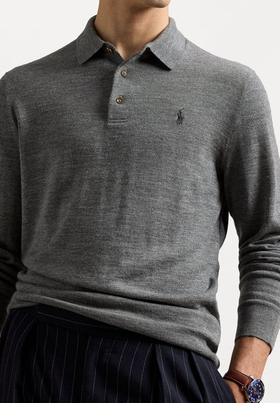 Pilkas megztinis su ilgomis rankovėmis ir apykakle, dviem mygtukais bei logotipu ant krūtinės. Minkšta tekstūra, derinama su tamsiai dryžuotomis kelnėmis.