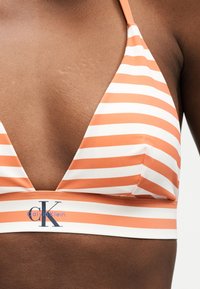 Nærbilde av en person som har på seg en oransje og hvitstripet Calvin Klein bikini-topp med dyp V-utringning.