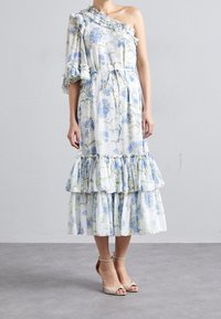 Robe midi florale, blanche avec des fleurs bleues, à une épaule avec volants, taille cintrée, volants en couches au bas, tissu doux, associée à des sandales à talons nus.
