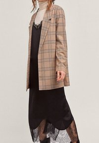 Béžový kostkovaný blazer přes černé šaty na ramínka s krajkovým lemem. Texturovaná látka, rovný střih, dlouhé rukávy s knoflíkovými detaily. Hladký, elegantní vzhled.