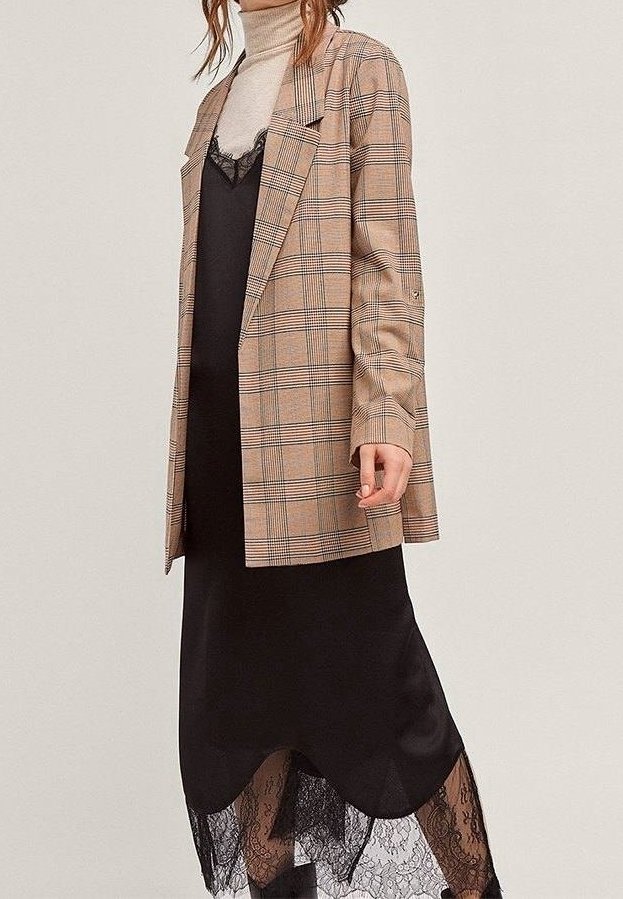 Béžový kostkovaný blazer přes černé šaty na ramínka s krajkovým lemem. Texturovaná látka, rovný střih, dlouhé rukávy s knoflíkovými detaily. Hladký, elegantní vzhled.